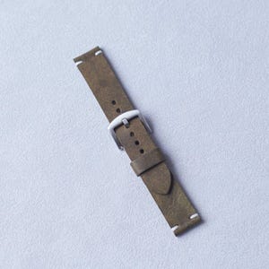 May include: Olive green leather watch strap with a silver-colored buckle. The strap has white stitching at the ends. The watch band is angled diagonally across a light gray surface.