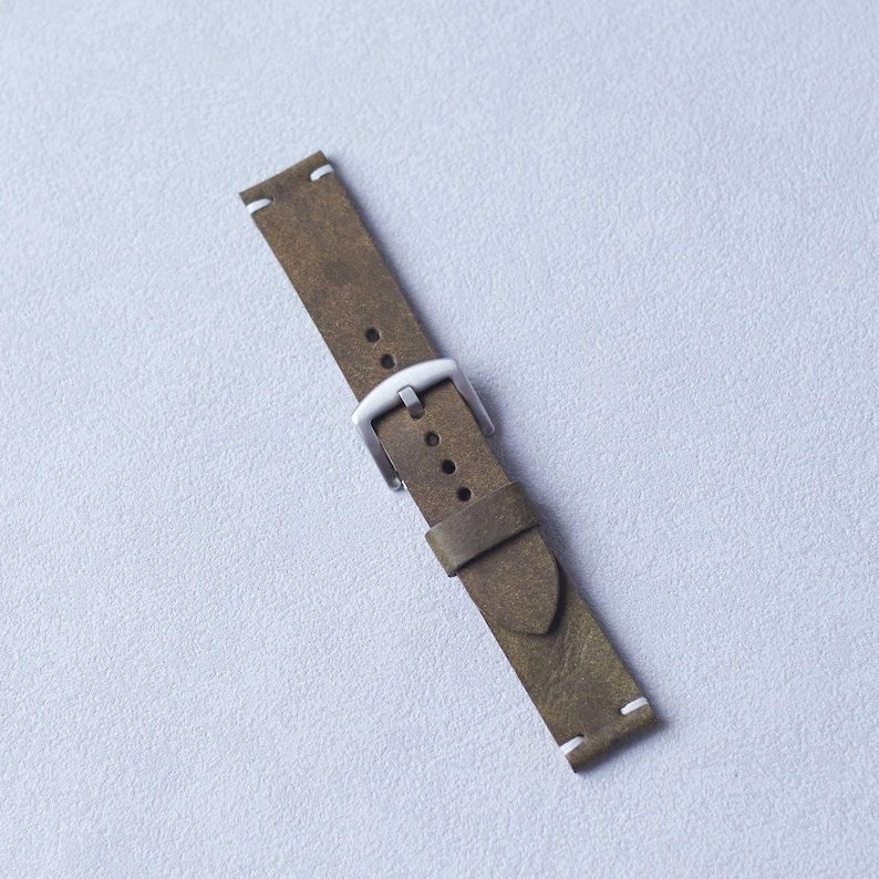 May include: Olive green leather watch strap with a silver-colored buckle. The strap has white stitching at the ends. The watch band is angled diagonally across a light gray surface.