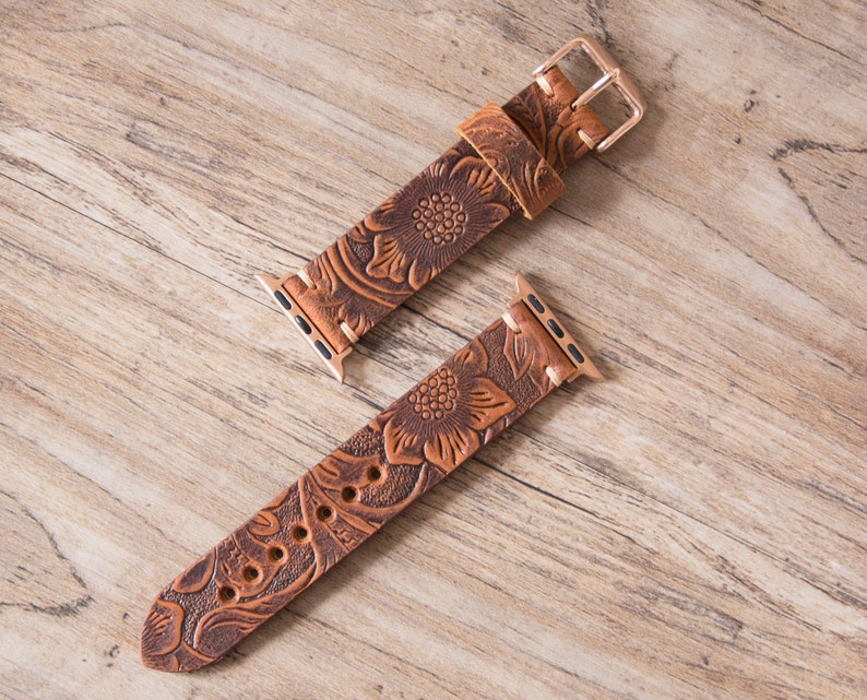 May include: Brown leather watch band with a floral embossed design. The band features a gold-colored buckle and connectors. The watch band is shown on a wooden surface.