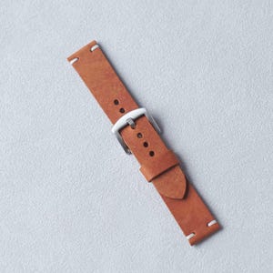 May include: A brown leather watch strap with a silver buckle and white stitching. The strap is rectangular with rounded edges and several holes for adjustment. The watch band is approximately 9 inches long.