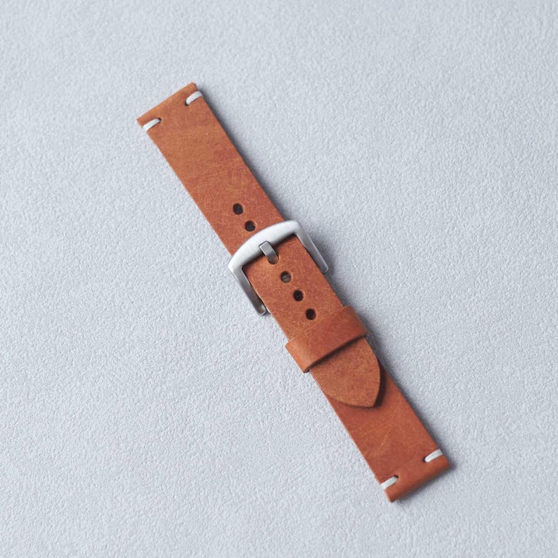 May include: A brown leather watch strap with a silver buckle and white stitching. The strap is rectangular with rounded edges and several holes for adjustment. The watch band is approximately 9 inches long.