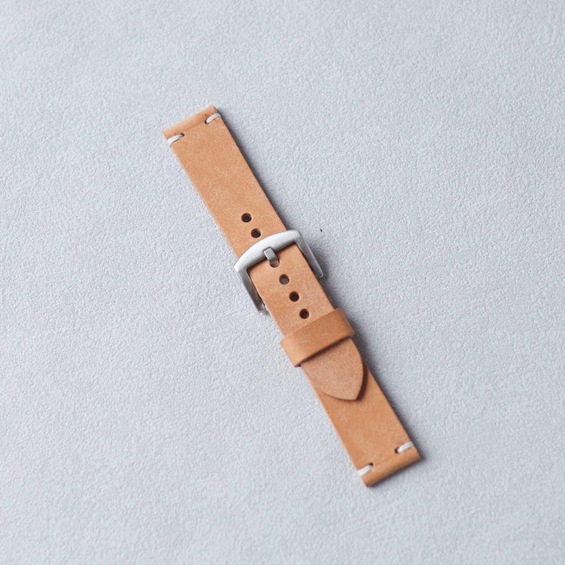 May include: A light brown leather watch strap with a silver buckle. The strap has white stitching at the ends and several holes for adjustment. The watch band is angled across a light gray surface.