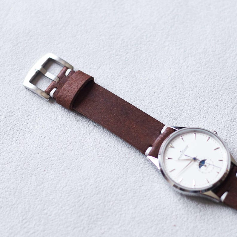 May include: A close-up of a wristwatch with a brown leather band and a silver buckle. The watch face is white with silver hands and a moon phase indicator. The band is stitched with white thread.
