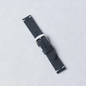 May include: A black leather watch strap with a silver buckle. The strap has white stitching at the ends. The watch band is angled on a light gray surface.