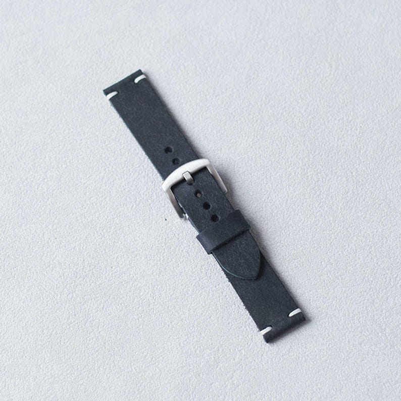 May include: A black leather watch strap with a silver buckle. The strap has white stitching at the ends. The watch band is angled on a light gray surface.