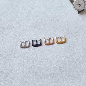 May include: Four watch buckles in silver, black, rose gold, and gold. The buckles are rectangular with a single bar. A wristwatch with a white face and a green band is in the background.