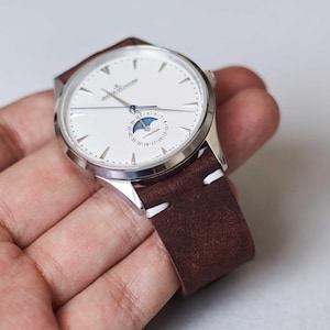 May include: A silver wristwatch with a white face, featuring a moon phase indicator and a brown leather strap with white stitching. The watch has a classic design and is held in a hand.