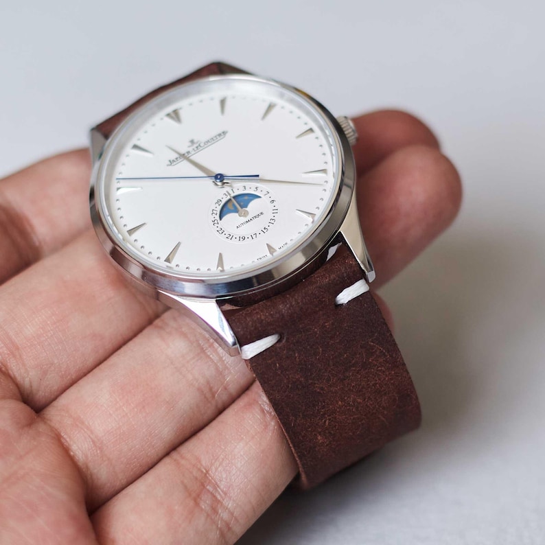 May include: A silver wristwatch with a white face, featuring a moon phase indicator and a brown leather strap with white stitching. The watch has a classic design and is held in a hand.