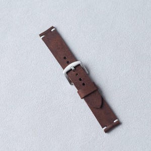 May include: A brown leather watch strap with a silver-colored buckle. The strap has white stitching at the ends and is designed to attach to a watch face. The leather has a textured appearance.