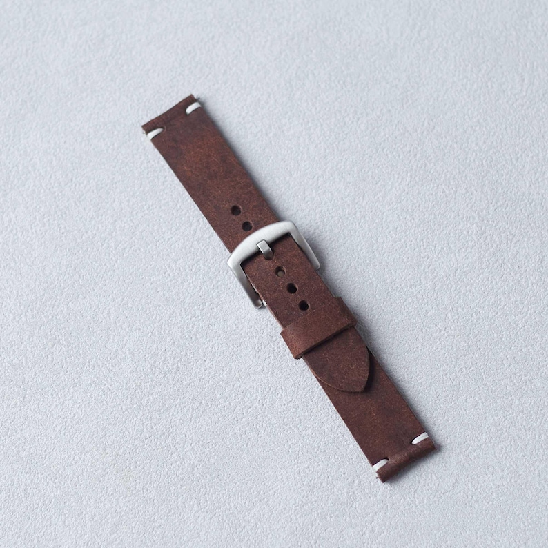 May include: A brown leather watch strap with a silver-colored buckle. The strap has white stitching at the ends and is designed to attach to a watch face. The leather has a textured appearance.