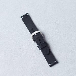 May include: A black leather watch strap with a silver-colored buckle. The strap has white stitching at the ends and is angled across a light gray surface. The watch band is a simple, classic design.