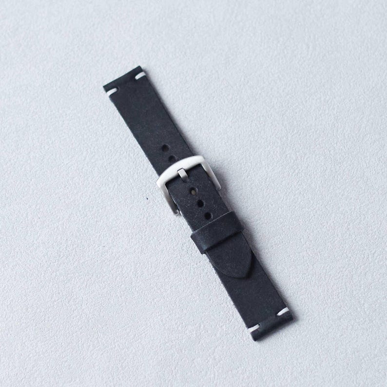 May include: A black leather watch strap with a silver-colored buckle. The strap has white stitching at the ends and is angled across a light gray surface. The watch band is a simple, classic design.