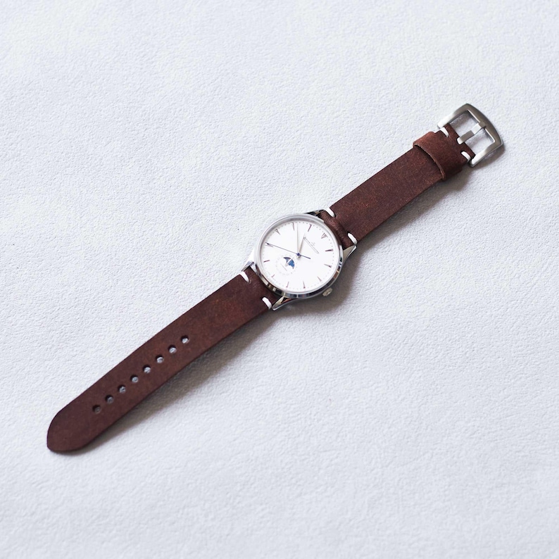 May include: A silver wristwatch with a white face and a brown leather band. The watch has a small moon phase indicator and white stitching. The band is approximately 0.75 inches wide and has a silver buckle.
