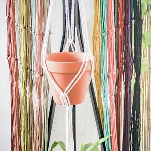 May include: A terracotta pot hangs from a macrame plant hanger made with colourful yarn strands. The hanger is attached to a black metal stand.