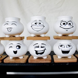May include: Eight white ceramic pots with different facial expressions. Each pot has three small feet and is sitting on a wooden base.