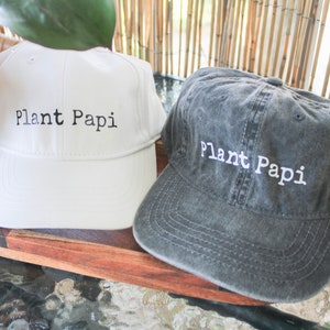 May include: Two baseball caps with the text "Plant Papi" embroidered on the front. One cap is white and the other is dark grey.