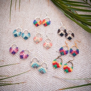 May include: A collection of 10 pairs of handmade heart-shaped earrings. The earrings are made with colorful thread and have silver hoops. The earrings are displayed on a light brown fabric background.