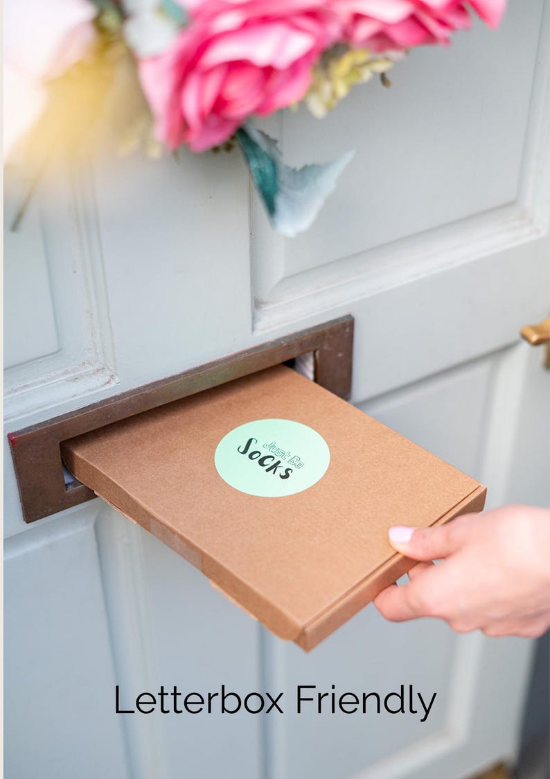 May include: A flat, rectangular brown cardboard shipping box being inserted into a metal letterbox slot. The box features a round, light green sticker with the text "Just Be SOCKS" printed in black. The box is designed to be letterbox friendly.