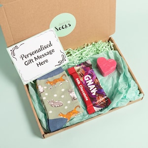 Könnte beinhalten: Eine braune Geschenkschachtel aus Karton mit einem Paar salbeigrünen Socken mit Katzenmuster, einer GNAW-Milchschokolade und einem rosa herzförmigen Gegenstand. Eine weiße Karte mit dem Text Personalised Gift Message Here liegt in der Schachtel.