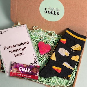 Op de afbeelding: Een kartonnen geschenkdoos met een paar zwarte sokken met een geel en wit kaaspatroon, een kleine Gnaw melkchocoladereep, een roze hartvormig item en een kaart met de tekst Personalised message here en Just Be Socks.