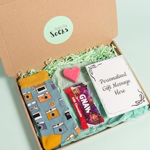 May include: A brown cardboard gift box containing a pair of blue socks with a camera pattern, a small pink heart-shaped item, a GNAW milk chocolate bar, and a white card with the text Personalised Gift Message Here, all on green tissue paper.