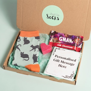May include: A cardboard gift box containing a pair of light blue socks with a dark gray cat pattern and orange cuffs, a bar of Gnaw milk chocolate, a small pink heart-shaped object, and a white card with the text Personalized Gift Message Here.
