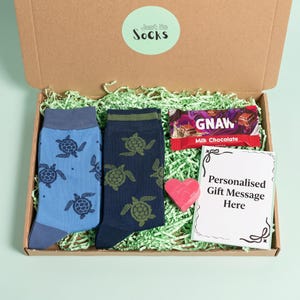 May include: A cardboard gift box containing two pairs of socks with turtle patterns, one light blue and one dark blue. Included is a Gnaw milk chocolate bar, a small pink heart-shaped object, and a card with the text Personalised Gift Message Here.