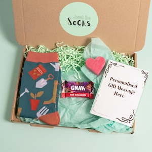 Könnte beinhalten: Eine Papp-Geschenkbox mit der Aufschrift Just Be Socks, die blaugrüne Socken mit Gartenmuster, eine rosa herzförmige Seife, einen Gnaw-Milchschokoladenriegel und eine weiße Karte mit dem Text Personalised Gift Message Here auf grünem Seidenpapier enthält.