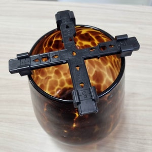 May include: Black plastic plant pot holder with a grid pattern. The holder is placed on top of a brown glass vase with a leopard print design.