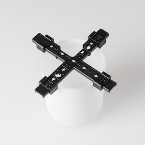 May include: A black plastic cross-shaped component, featuring small holes and rectangular tabs. It is placed over a white, rounded container. The design implies a structural or organizational function, possibly for a DIY project.