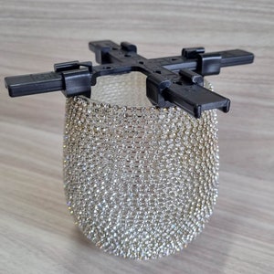 May include: A black plastic frame with four adjustable arms holds a round, clear glass vase covered in a sparkling, silver rhinestone mesh.