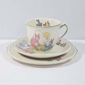 Könnte beinhalten: Ein Vintage-Kinder-Tee-Set mit weißem Hintergrund und grünem Rand. Tasse und Untertasse zeigen eine fantasievolle Illustration von Hasen und Kindern beim Spielen.