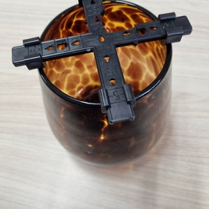 Puede incluir: Soporte de planta de plástico negro con cuatro brazos, diseñado para encajar en la parte superior de un jarrón o recipiente de vidrio redondo. El jarrón tiene un patrón de estampado de leopardo marrón y beige.