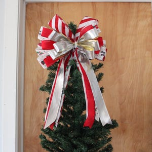 May include: A festive Christmas tree topper featuring a large bow with red and white striped ribbons, silver ribbons, and a gold accent. The bow is atop a small, green artificial Christmas tree.