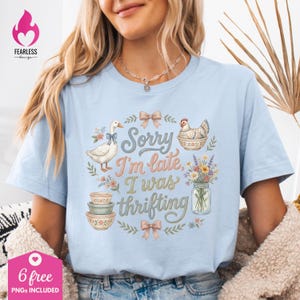 May include: Light blue t-shirt with the text "Sorry I'm late, I was thrifting." The design includes a goose, a chicken, flowers, and bowls. The shirt has a crew neck and short sleeves. The Fearless Design logo is in the upper left corner.