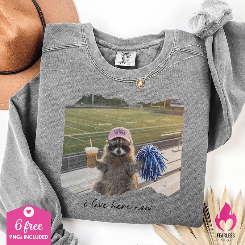 Puede incluir: Una sudadera gris de cuello redondo con un gr&aacute;fico de un mapache con una gorra rosa de Football Mom y gafas de sol, sosteniendo un caf&eacute; helado y un pomp&oacute;n azul y blanco. El texto debajo de la imagen dice, i live here now.