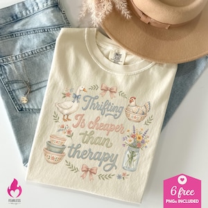 Puede incluir: Camiseta color crema con la frase "Thrifting Is cheaper than therapy" y dibujos de un ganso, una gallina, cuencos y flores. La camiseta est&aacute; colocada sobre unos vaqueros azules. Tambi&eacute;n se ven un sombrero y un collar.