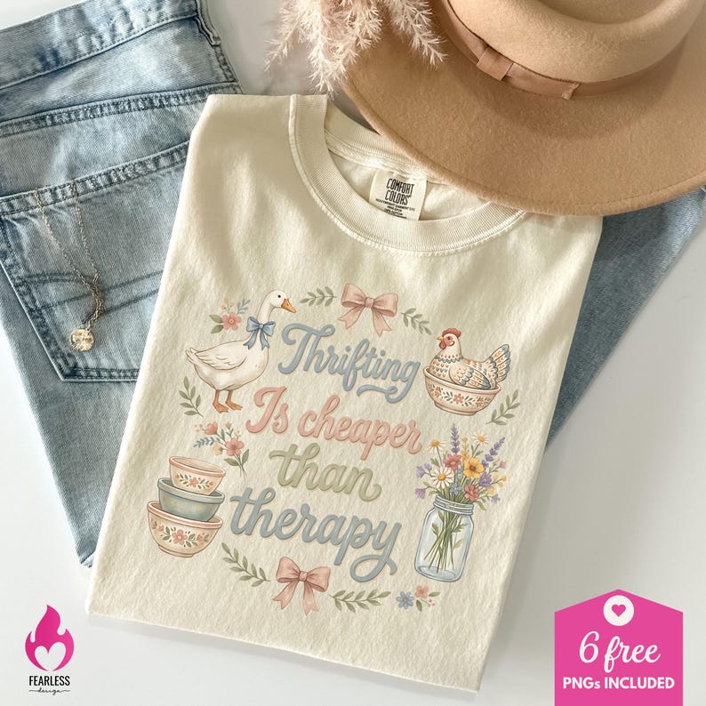 Puede incluir: Camiseta color crema con la frase "Thrifting Is cheaper than therapy" y dibujos de un ganso, una gallina, cuencos y flores. La camiseta est&aacute; colocada sobre unos vaqueros azules. Tambi&eacute;n se ven un sombrero y un collar.
