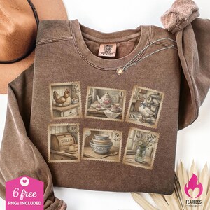 May include: Brown Comfort Colors sweatshirt featuring postage stamp designs with illustrations of a hen, teapot, goose, bread, bowls, and flowers. A silver necklace and a brown hat are also visible.
