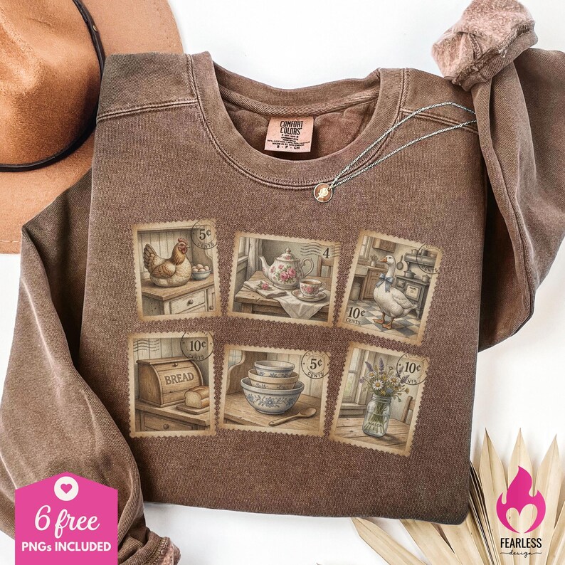 May include: Brown Comfort Colors sweatshirt featuring postage stamp designs with illustrations of a hen, teapot, goose, bread, bowls, and flowers. A silver necklace and a brown hat are also visible.