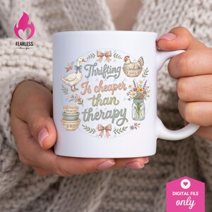 Puede incluir: Taza de cer&aacute;mica blanca con el texto "Thrifting Is cheaper than therapy", rodeada de ilustraciones de un ganso, una gallina, flores y cuencos. La taza tiene un asa curva y est&aacute; sujeta por una persona.