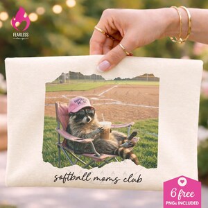 Può includere: Borsa tote color crema con un'illustrazione di procione con occhiali da sole e cappello rosa, che tiene in mano una bevanda. L'immagine è ambientata su uno sfondo di campo da baseball, con la scritta "softball moms club".