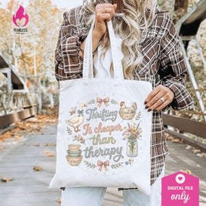 Puede incluir: Una bolsa de mano blanca con el texto "Thrifting Is cheaper than therapy" en un dise&ntilde;o circular. El dise&ntilde;o incluye ilustraciones de un ganso, una gallina, cuencos, flores y lazos. La bolsa la sostiene una persona que lleva una camisa a cuadros.