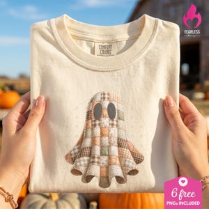 May include: A cream-colored short-sleeve t-shirt featuring a central graphic of a ghost composed of various brown, tan, and sage green plaid and floral fabric patches. The ghost has two dark oval eyes. The shirt has a Comfort Colors brand label.