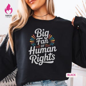 May include: Black sweatshirt with the text "Big Fan of Human Rights" in white, with colorful accents. The sweatshirt has long sleeves and a crew neck. The design includes a rainbow-colored burst.