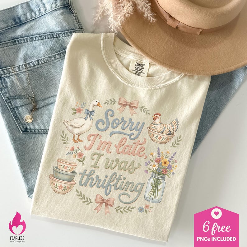 May include: Cream-colored t-shirt with the text "Sorry I'm late, I was thrifting" in pastel colors. The shirt features illustrations of a goose, a chicken, bowls, and flowers. The shirt is laid on top of blue jeans.