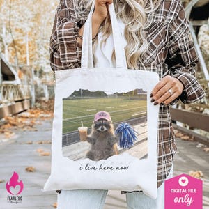 Puede incluir: Una bolsa de tela blanca con una fotograf&iacute;a impresa de un mapache que lleva un gorro rosa con la inscripci&oacute;n Football Mom, sosteniendo un caf&eacute; y un pomp&oacute;n azul y blanco en un campo de f&uacute;tbol, con el texto i live here now impreso debajo.