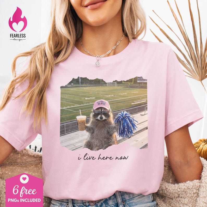 Puede incluir: Una camiseta rosa claro con el gr&aacute;fico de un mapache que lleva una gorra rosa y gafas, sosteniendo una bebida y un pomp&oacute;n azul y blanco en un campo de f&uacute;tbol. El texto "i live here now" est&aacute; impreso en negro debajo de la imagen.
