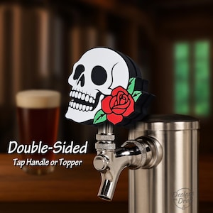 May include: A double-sided tap handle or topper featuring a white skull with black accents and a red rose. The handle is mounted on a stainless steel tap.