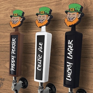 Pode incluir: Três puxadores de torneira de cerveja com um design de duende. As pegas são castanhas, brancas e pretas, cada uma com um tipo de cerveja diferente: Paddy Porter, Celtic Ale e Lucky Lager. Montadas numa superfície de madeira.